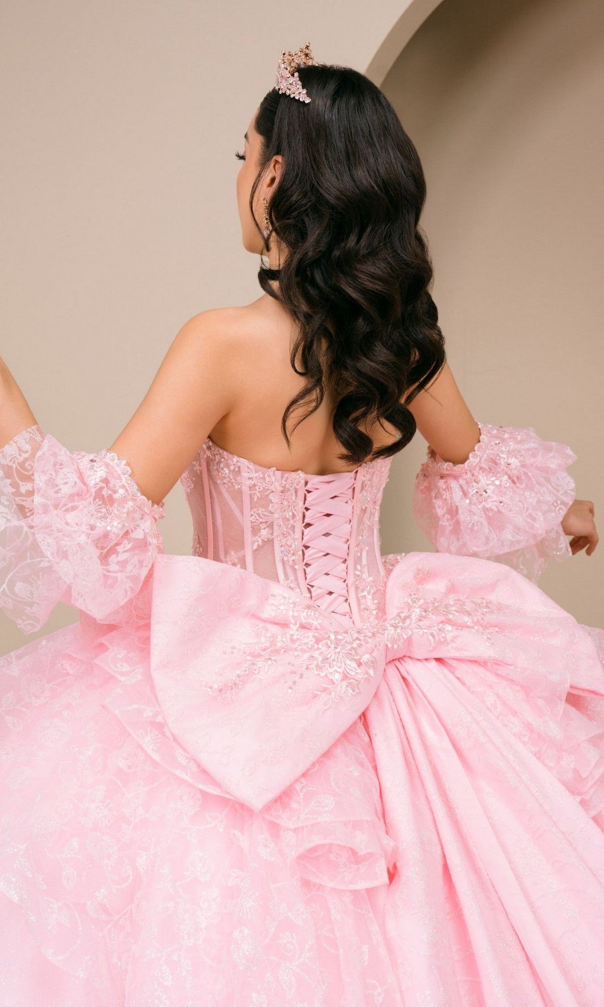 A woman in the Quinceanera Dress 8535JQ by Cinderella Coutureโa pink ball gown featuring lace sleeves and a large bow on the backโstands indoors facing away from the camera, wearing a tiara.